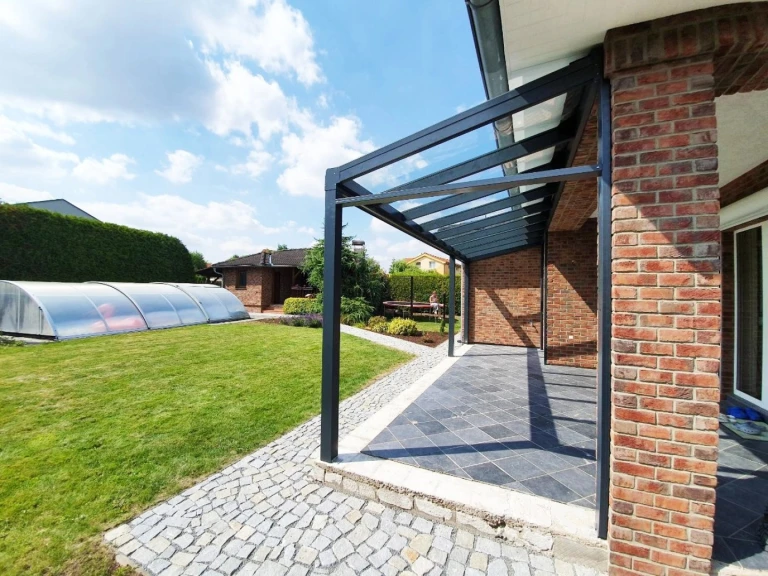 A modern anthracite-colored aluminum pergola with a glass roof next to a brick house with a garden and a pool.