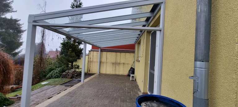 A modern gray aluminum LIGHT pergola with a glass roof, mounted against the wall of the house above the patio