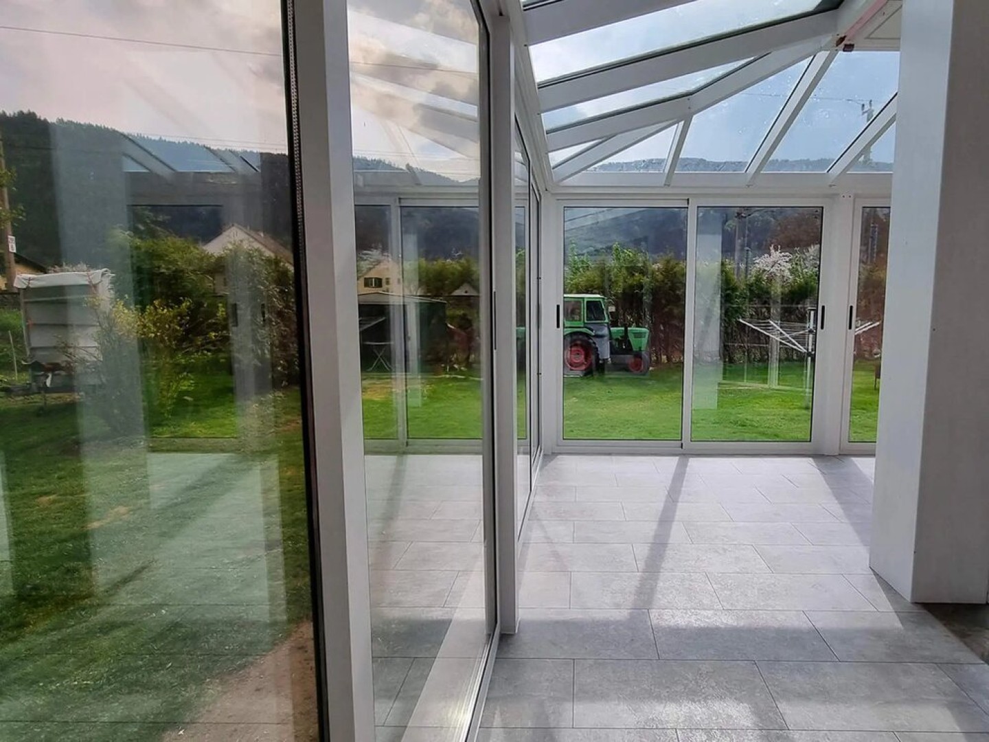 A view from inside the house of the glass-enclosed terrace with a garden and a tractor in the background