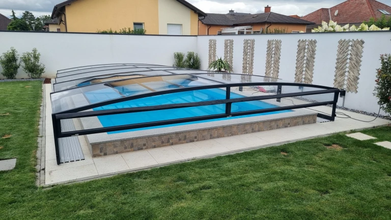 A modern, low-profile pool enclosure with a black frame and transparent panels in the garden