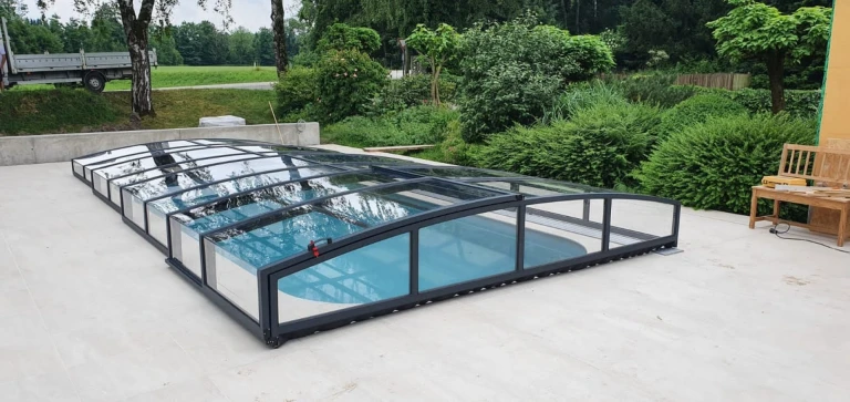 A modern, low-profile pool enclosure with an anthracite frame and clear panels in the garden