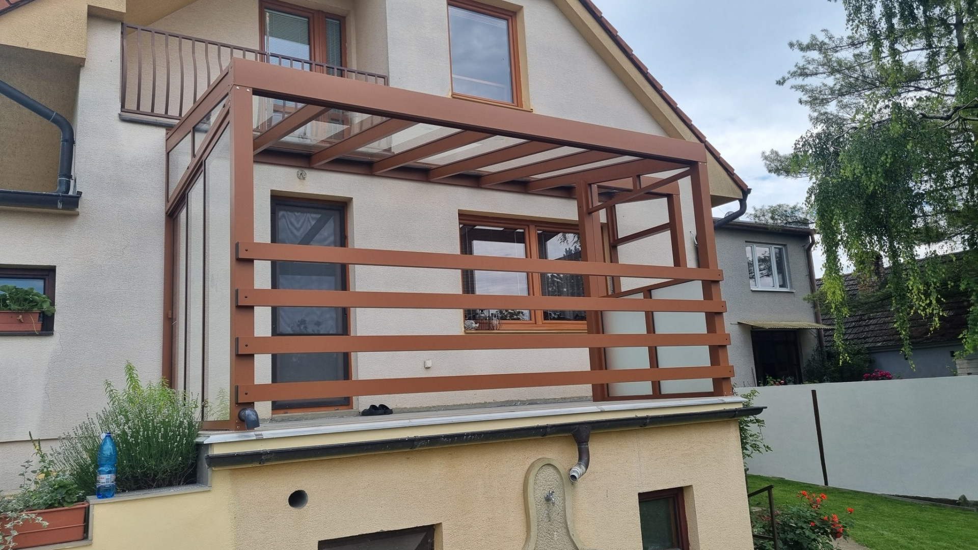 A brown, custom-designed aluminum pergola with a glass roof and side shading on the balcony of a single-family home