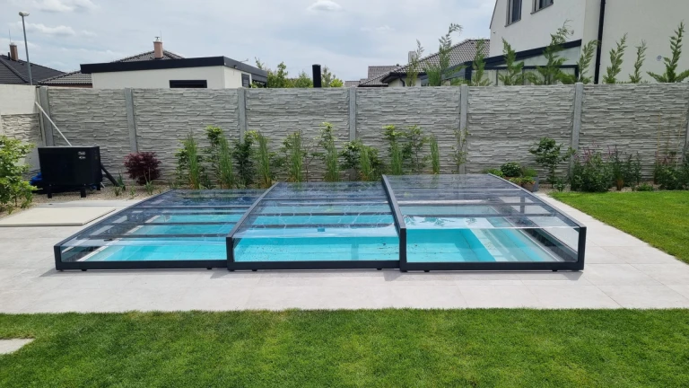 A modern, low-profile pool enclosure with an anthracite-colored frame and clear panels in the garden of a single-family home.