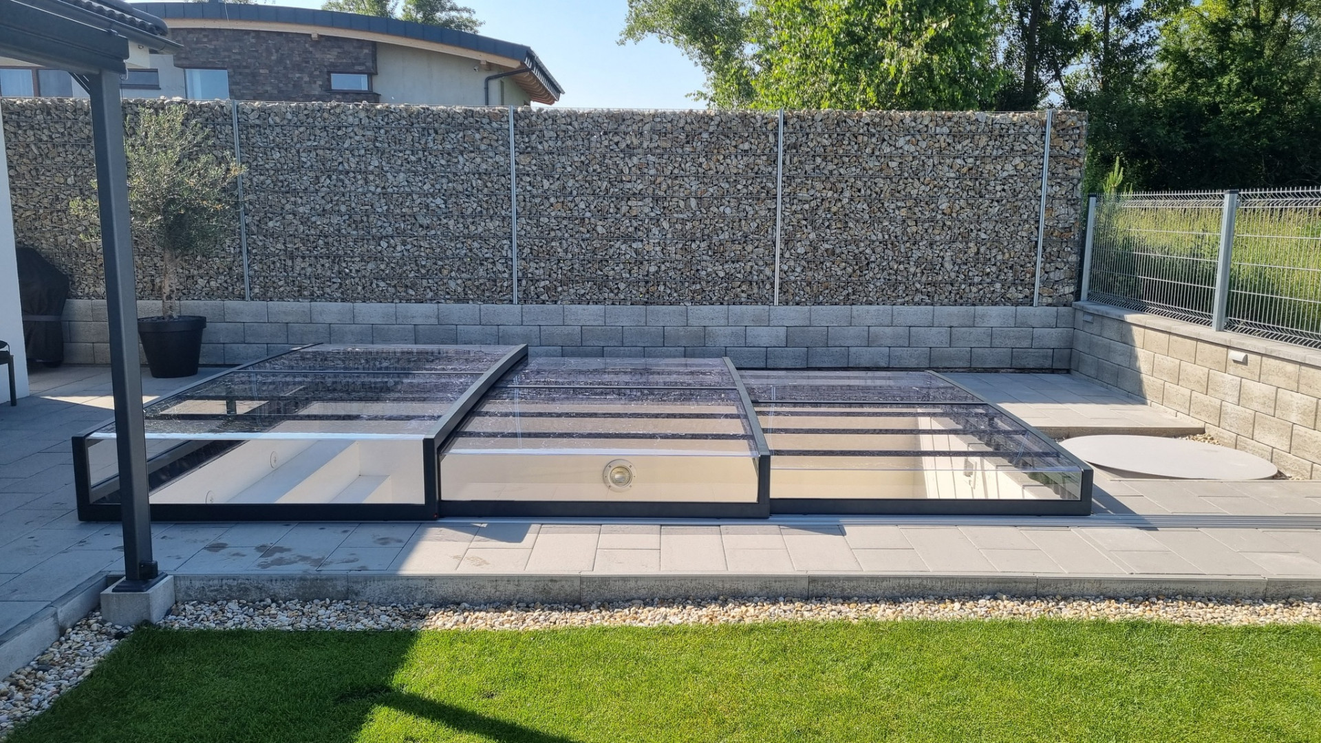 A low-profile EXCLUSIVE pool enclosure made of clear polycarbonate in the garden next to a stone wall.