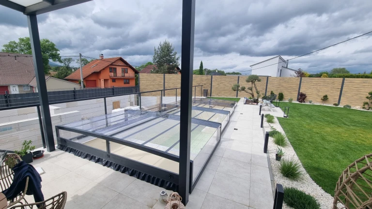 A view of a modern, low-slung pool canopy in a landscaped garden under a cloudy sky.