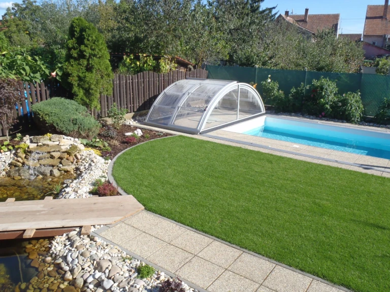 A modern, low-profile KLASIK pool enclosure with light blue water and a landscaped garden featuring a small pond.