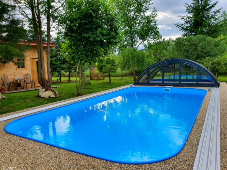 A blue in-ground pool in the garden with a retractable canopy and a wooden cottage in the background