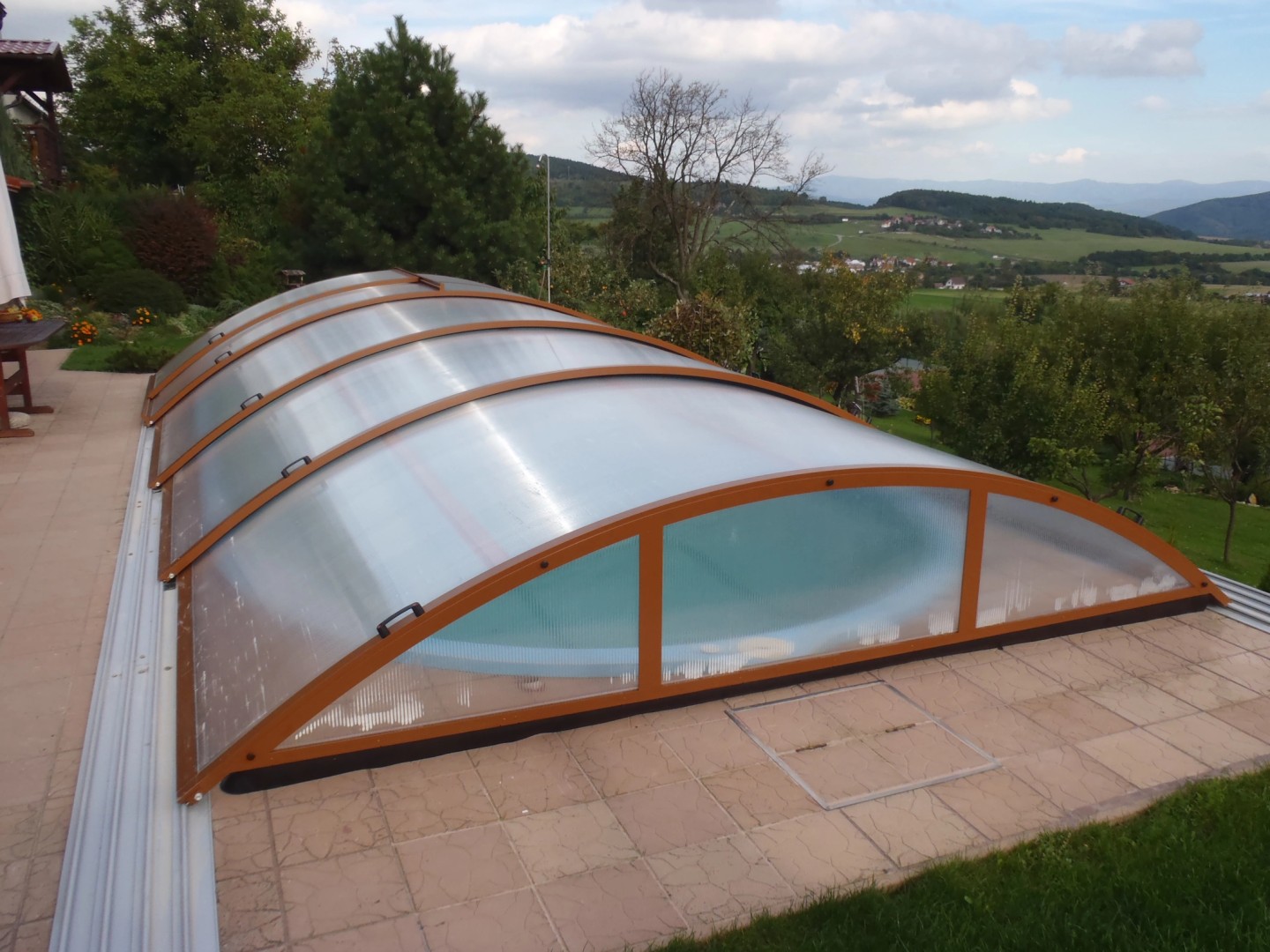 A closed, low-arched pool canopy with a brown frame, located on a terrace overlooking the hilly landscape.