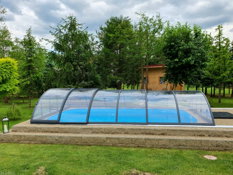 A closed, low, arched pool cover over a blue pool in a garden with trees.
