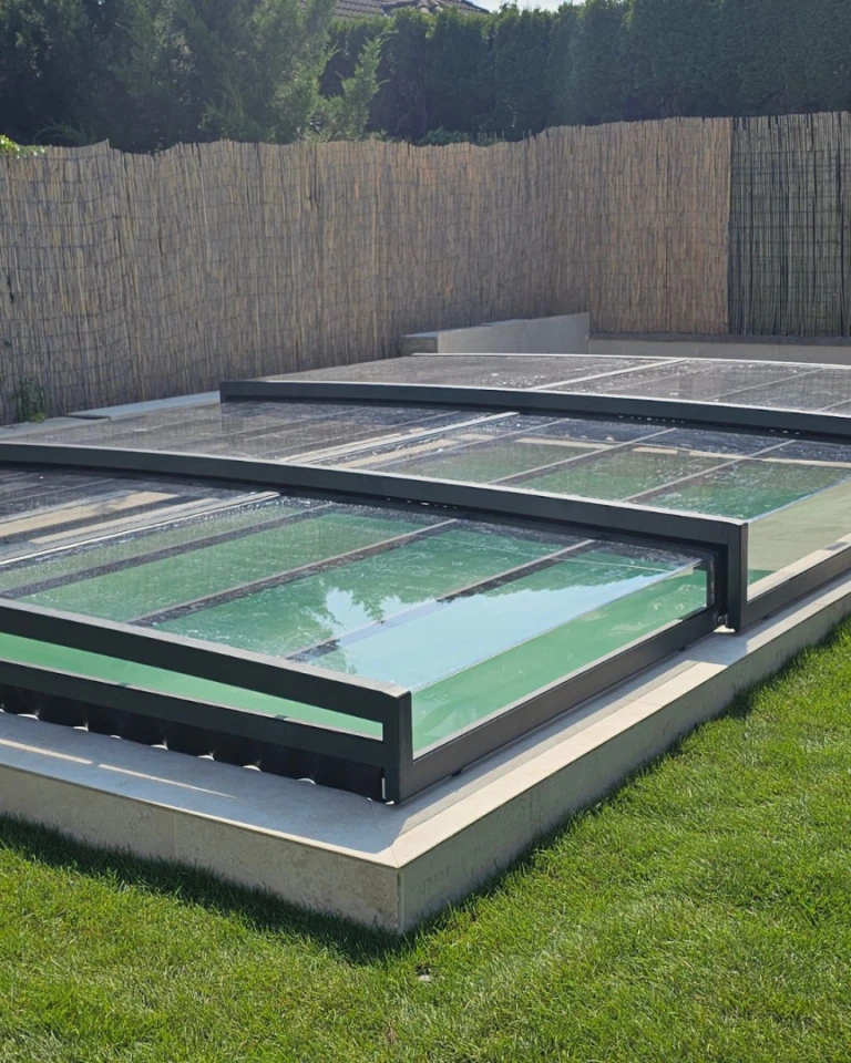 A modern, low-profile pool enclosure with an anthracite frame and clear panels in the garden