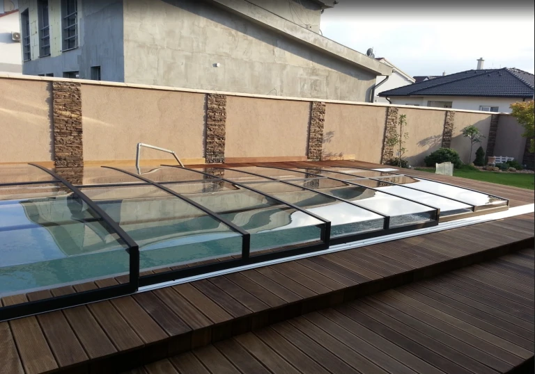 A modern, low-profile pool enclosure with a black frame and clear panels on a wooden-decked terrace.