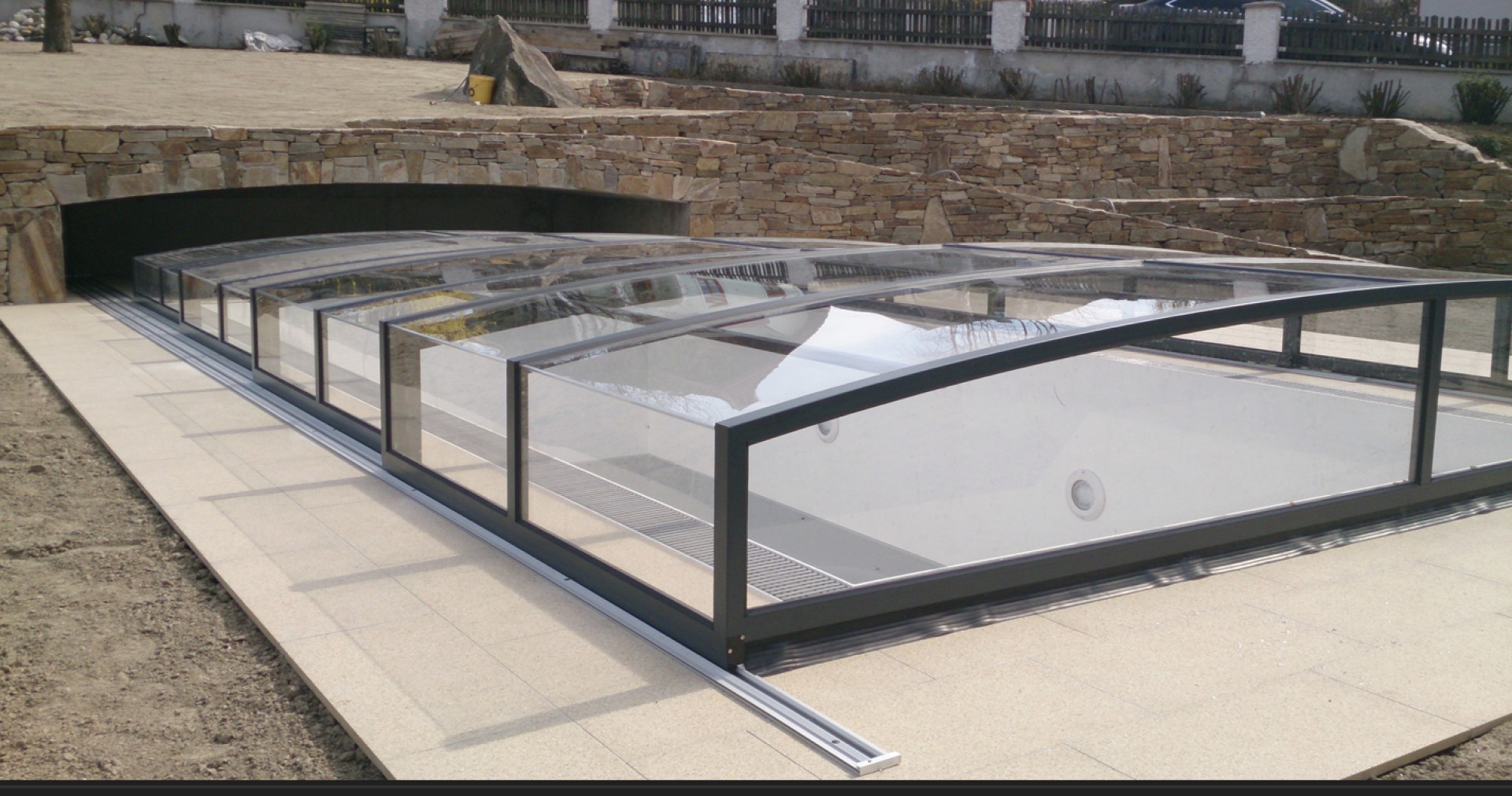 A modern, low-profile pool enclosure with transparent panels in a garden with stone paving.