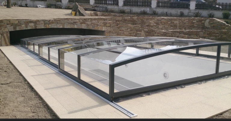 A modern, low-profile pool enclosure with clear walls set against a stone exterior