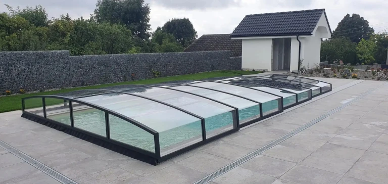 A modern, low-profile pool enclosure with a black frame and clear glass on the terrace next to the house.