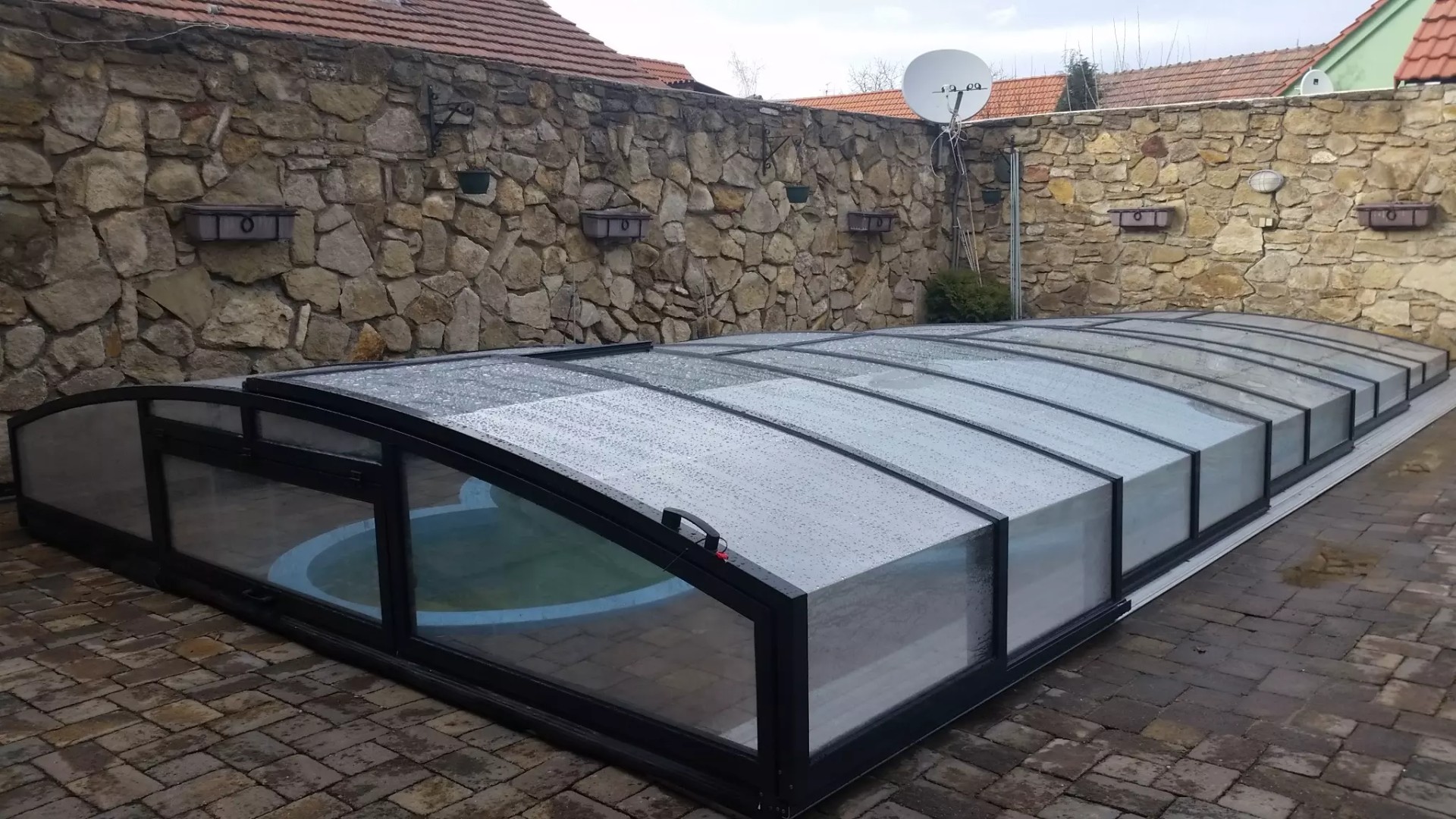 A low-profile pool enclosure with dark-colored framing in a backyard with a stone wall.