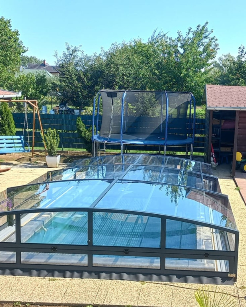 A modern, low-profile pool enclosure with an anthracite frame in a garden featuring a trampoline