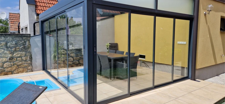 A modern, glass-enclosed sunroom with an anthracite-colored frame, built next to the pool by the house.