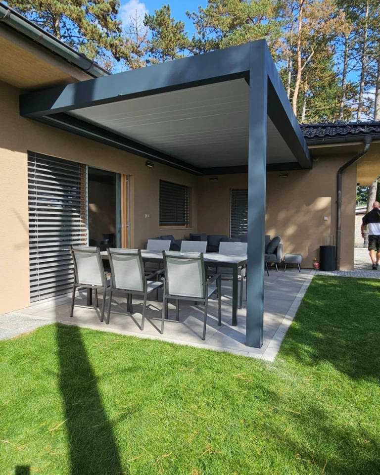 A modern gray bioclimatic pergola next to the house, with garden furniture and a lawn