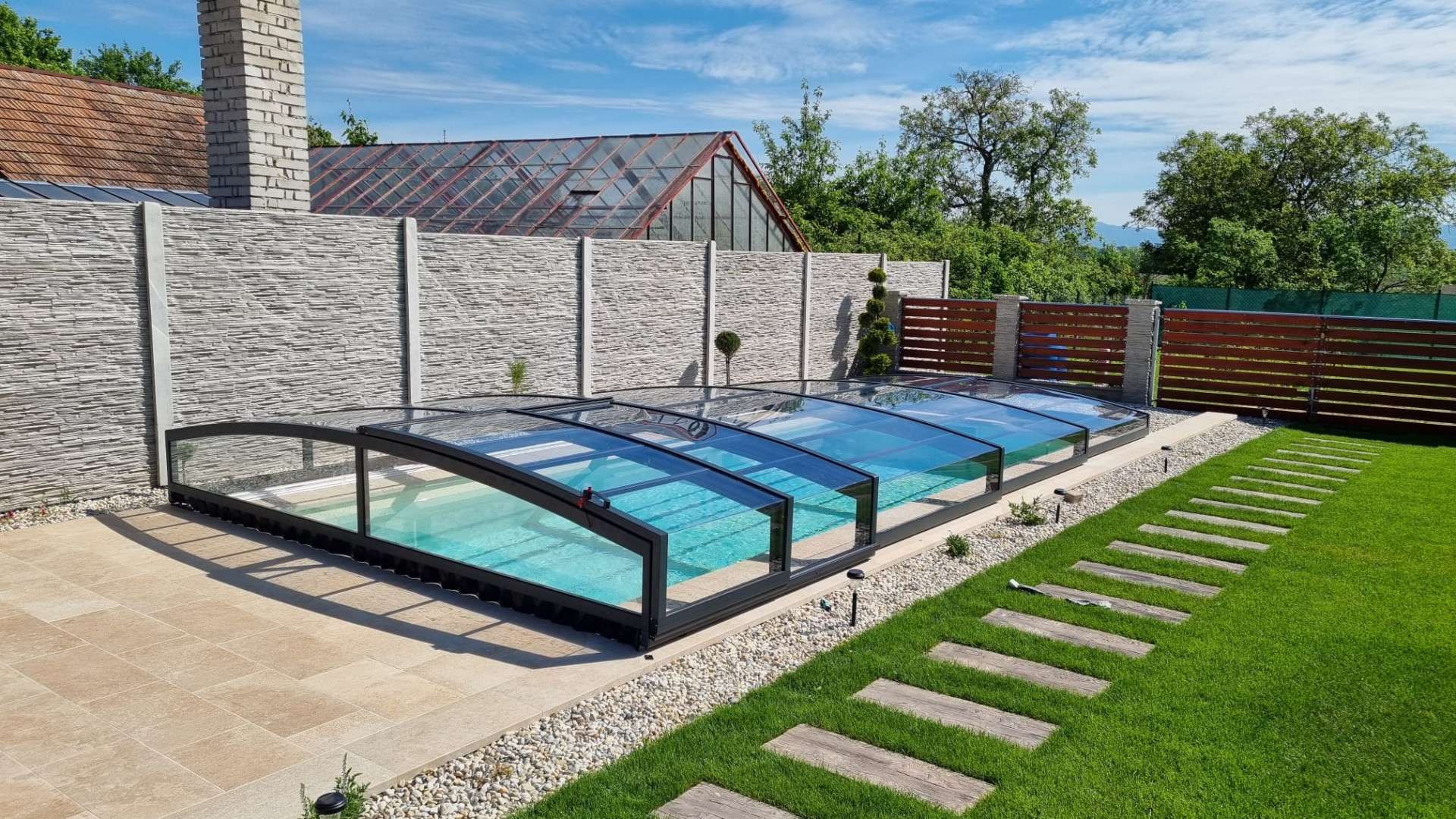 A modern, low-profile pool enclosure with an anthracite frame and clear glass in a well-maintained garden.