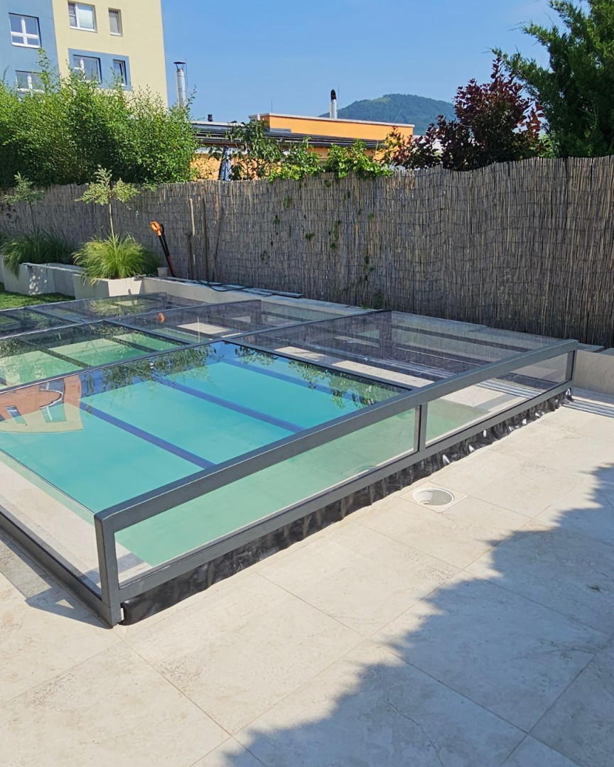 A modern, low-profile glass pool enclosure with an anthracite frame on a paved terrace.