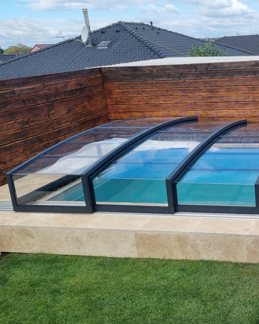 A modern, low-profile pool enclosure with an anthracite frame and clear paneling next to a wooden fence