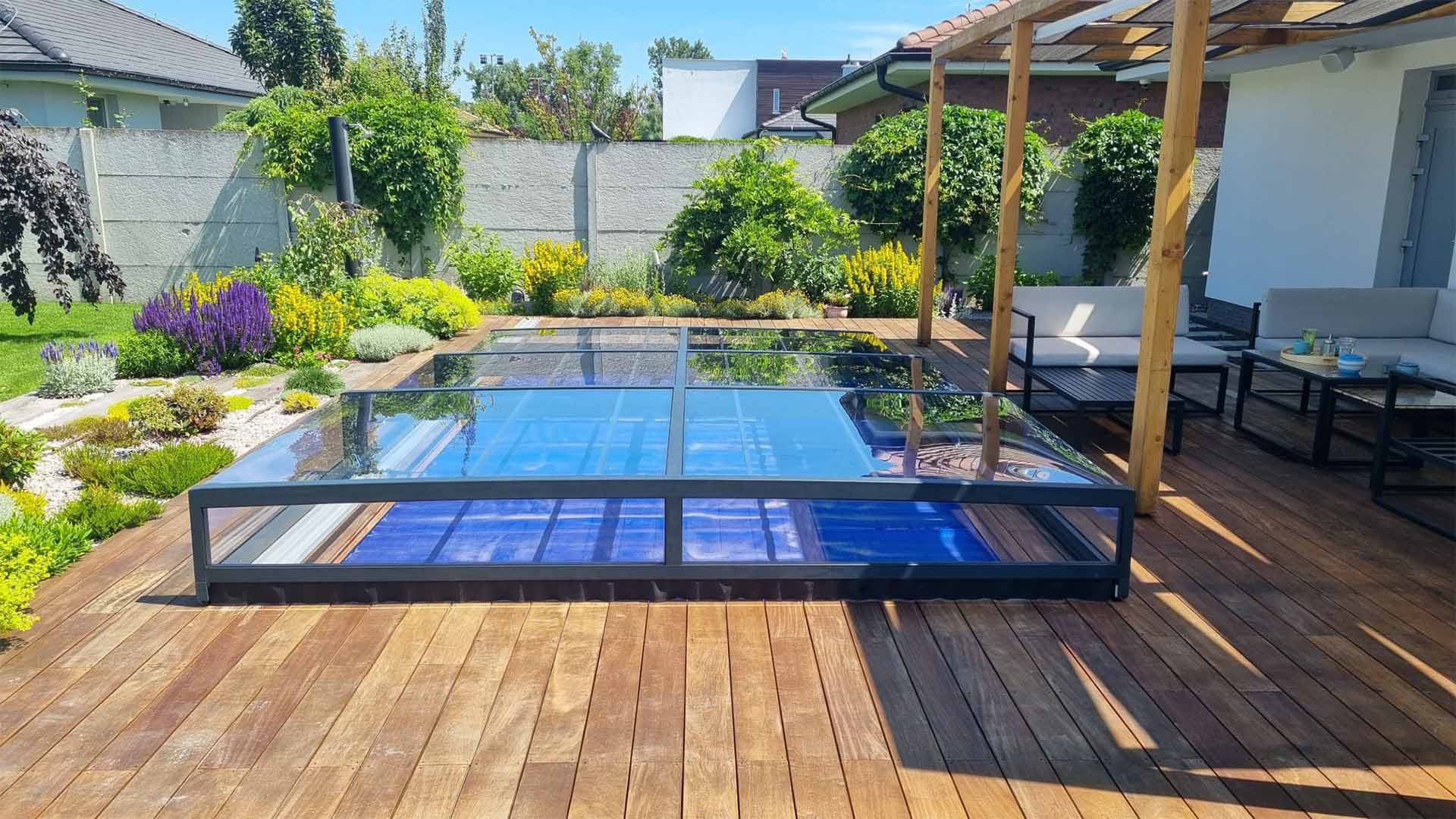 A modern, low-profile glass pool enclosure on a wooden deck in the garden
