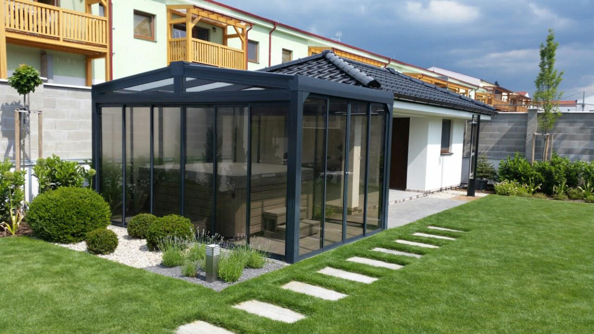 A modern, glass-enclosed hot tub shelter with a dark frame in the landscaped garden next to the house.