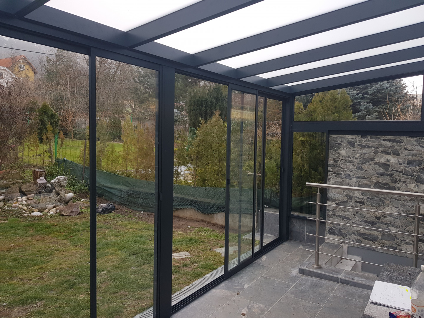 A modern, glass-enclosed sunroom with an aluminum frame and sliding doors offering a view of the garden