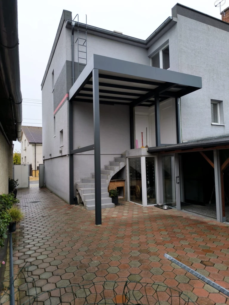 A modern dark gray aluminum pergola with a fixed roof attached to the facade of a single-family home above the outdoor staircase.