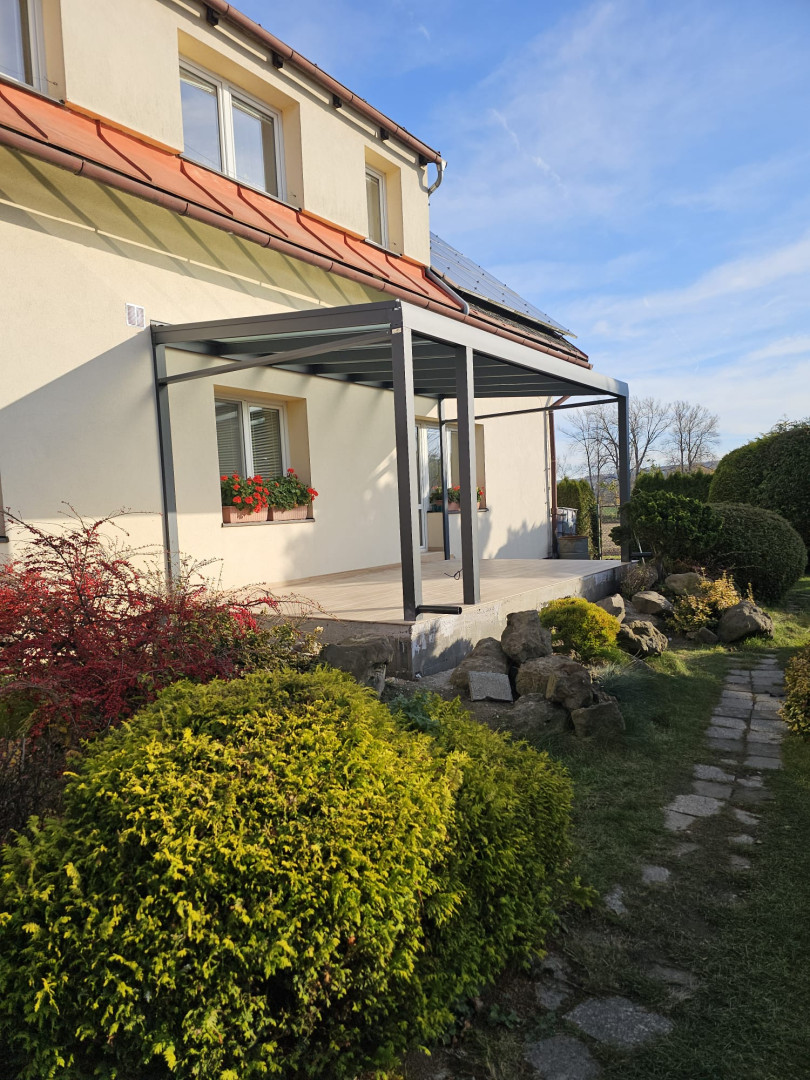 A modern aluminum pergola with a glass roof attached to a single-family home with a garden.