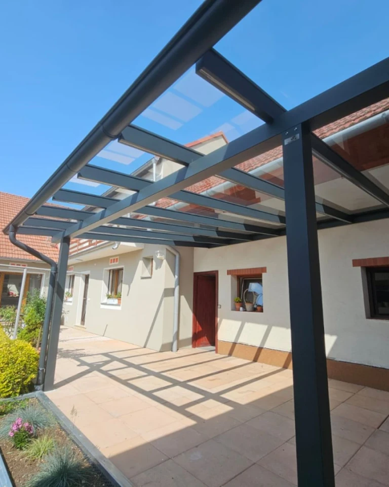 A modern anthracite-colored aluminum pergola with a glass roof attached to a single-family home with a paved patio.