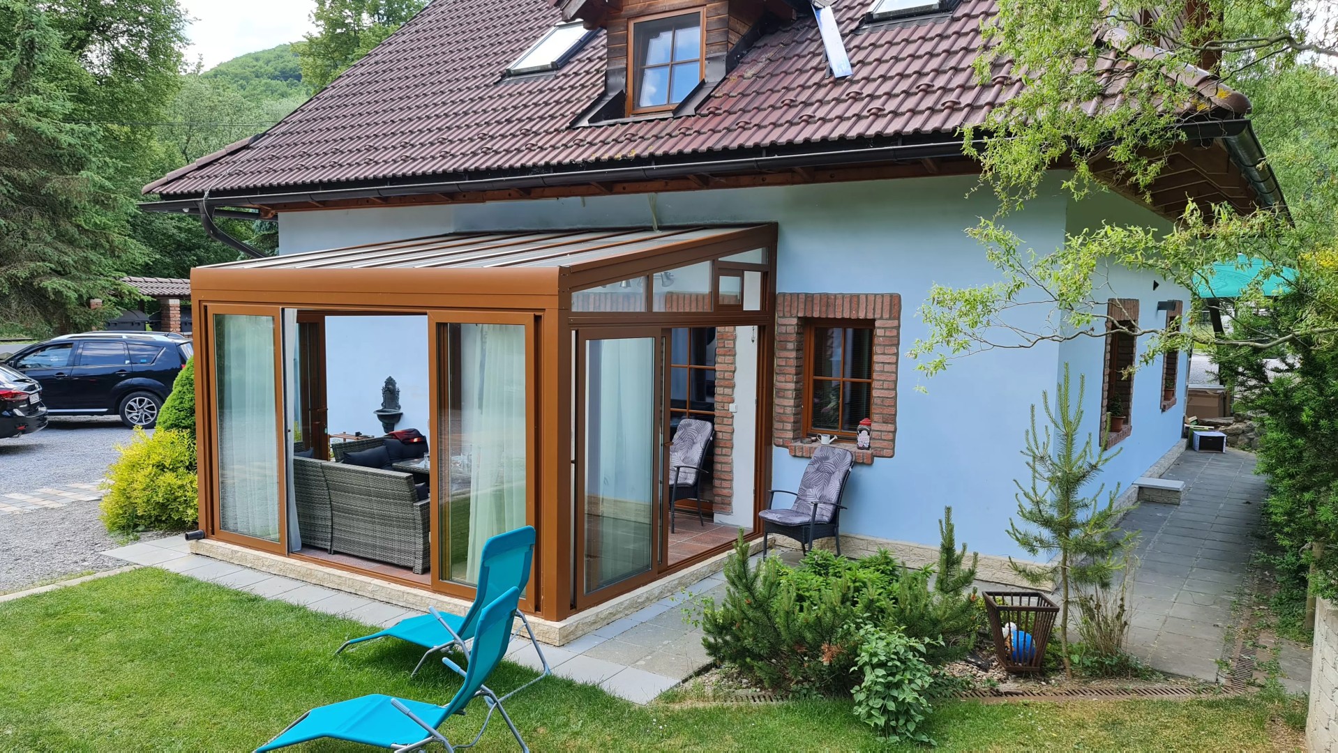 A brown ROBUST aluminum pergola with glass panels attached to a single-family home, complete with garden furniture.