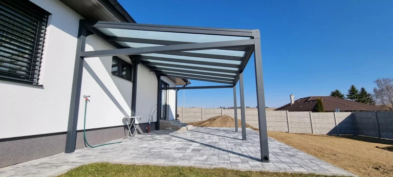 A modern, anthracite-colored ROBUST aluminum pergola with a glass roof installed next to a single-family home in Jihlava.