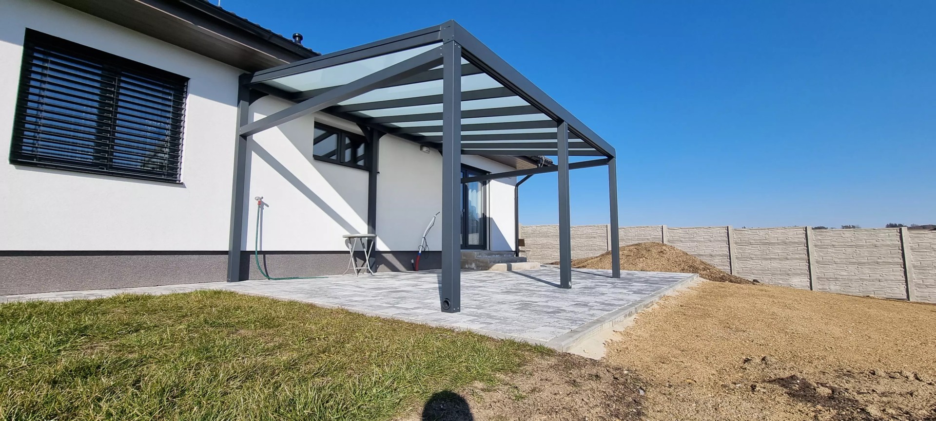 A modern anthracite-colored aluminum pergola with a glass roof, attached to a single-family home on a tiled patio.