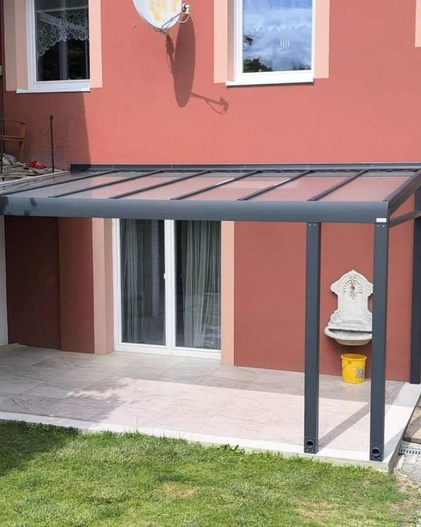 A modern aluminum pergola with a glass roof, attached to the wall of the red house above the patio.
