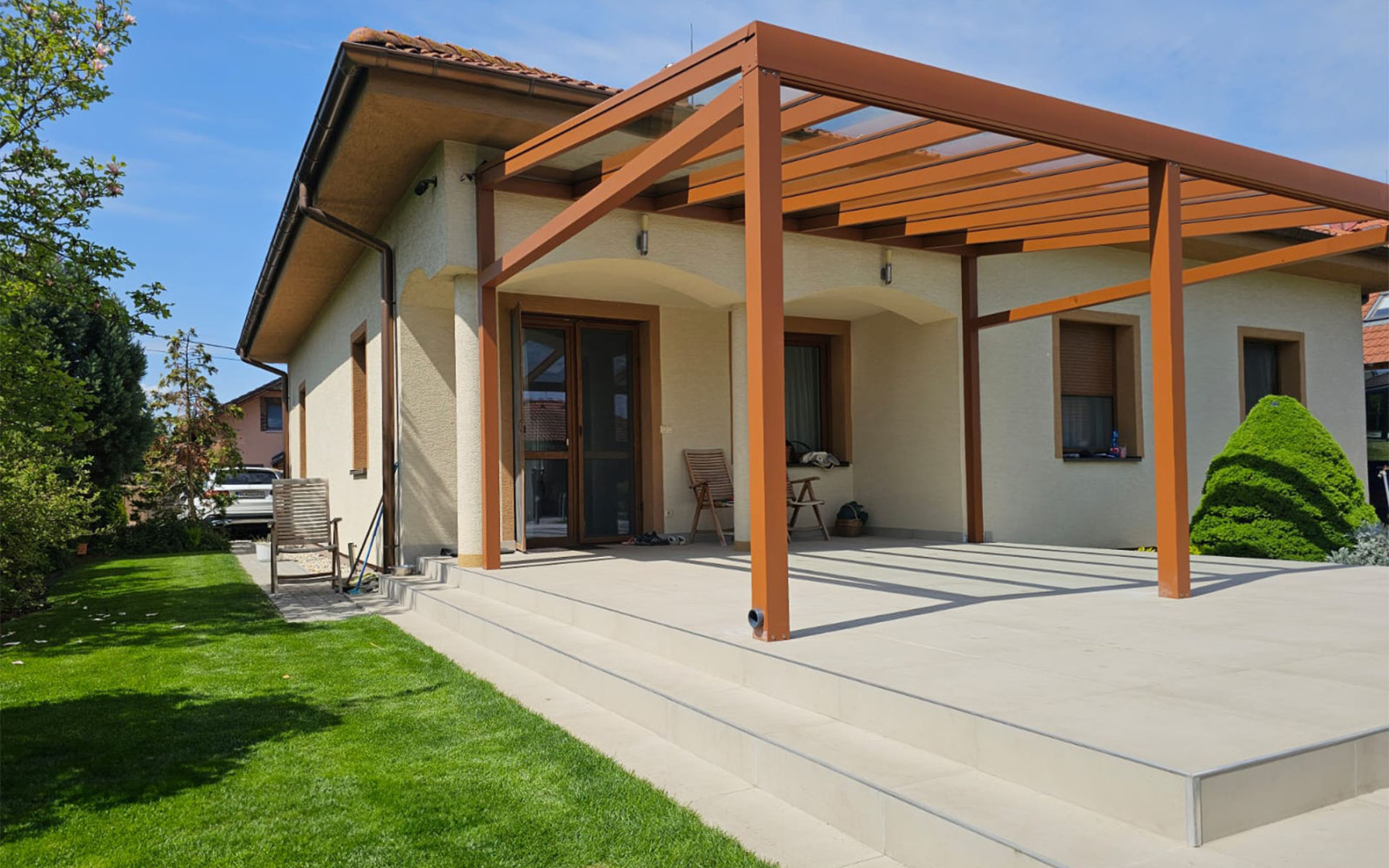A modern brown aluminum pergola with a glass roof attached to a single-family home with a spacious terrace.