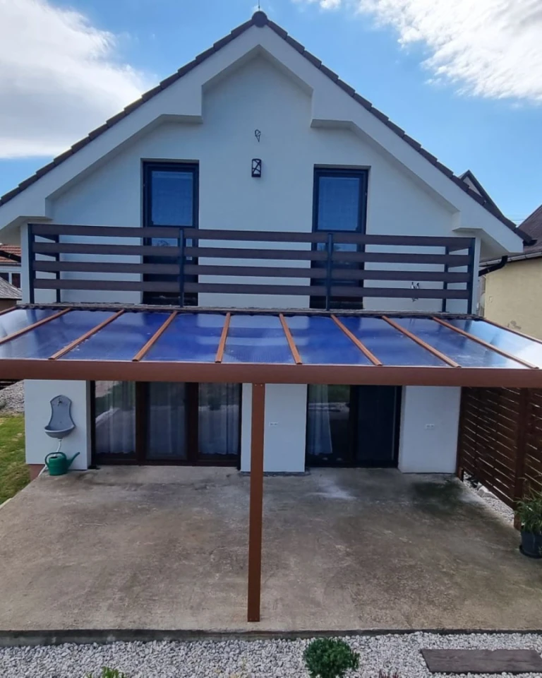 A brown aluminum pergola with a glass roof attached to a single-family home with a balcony.