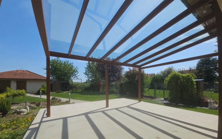 A view from under a glass-enclosed aluminum pergola of the garden with a single-family home