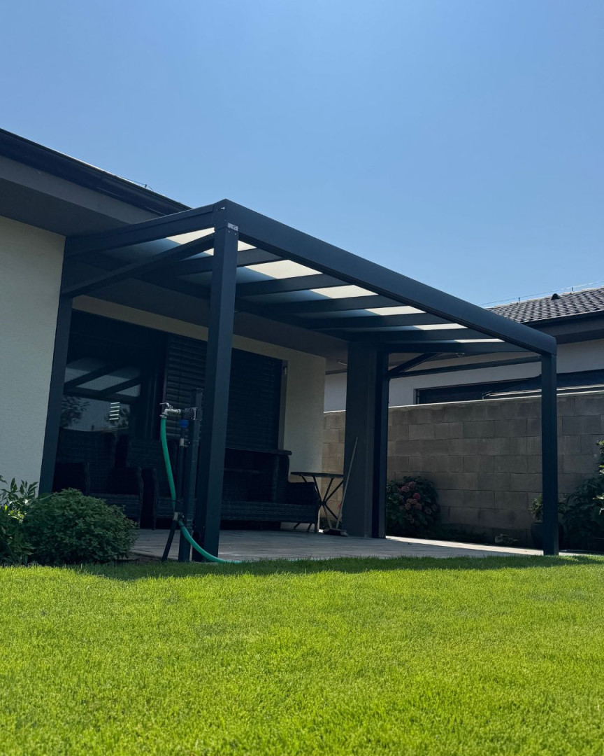 A modern dark-gray aluminum pergola attached to the house, with a glass roof and a manicured lawn