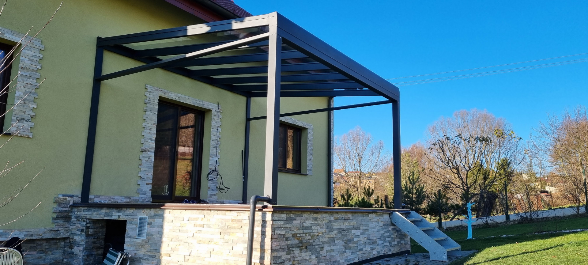 A modern, dark gray ROBUST aluminum pergola with a glass roof installed on the patio of a single-family home.