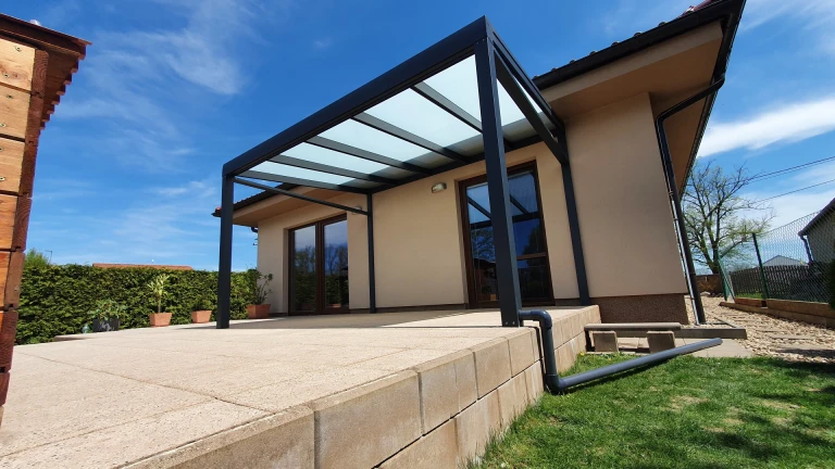 A modern dark gray aluminum pergola with a glass roof, installed on a paved patio attached to a single-family home.