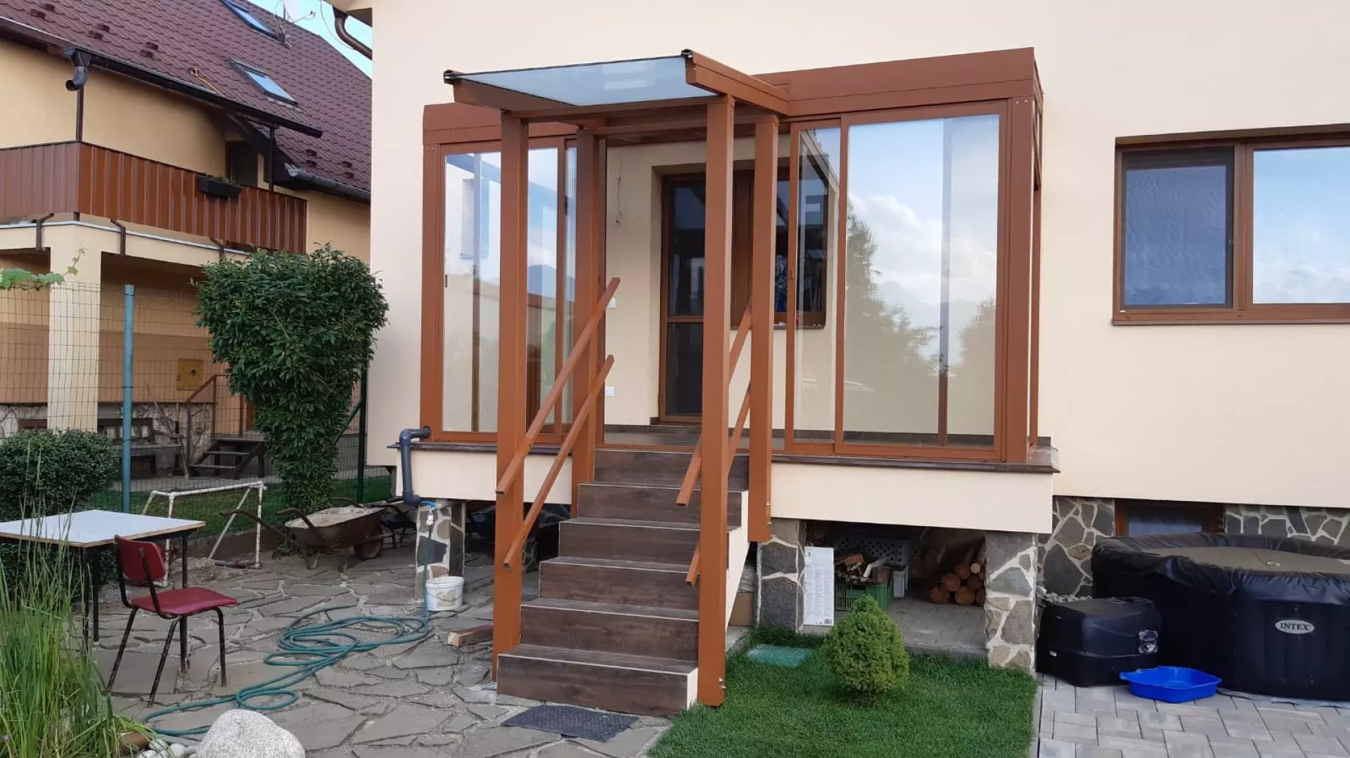 A brown, glass-enclosed aluminum pergola with a canopy over the entrance stairs of a single-family home.