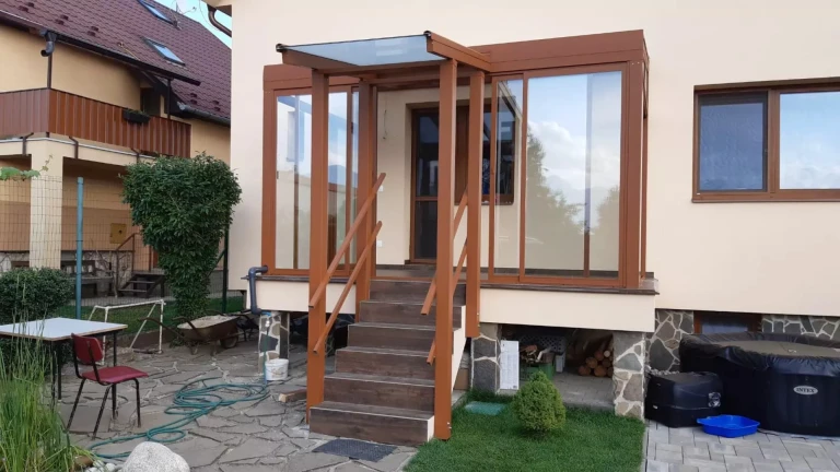 A brown aluminum pergola with glass panels and a canopy over the staircase at a single-family home.