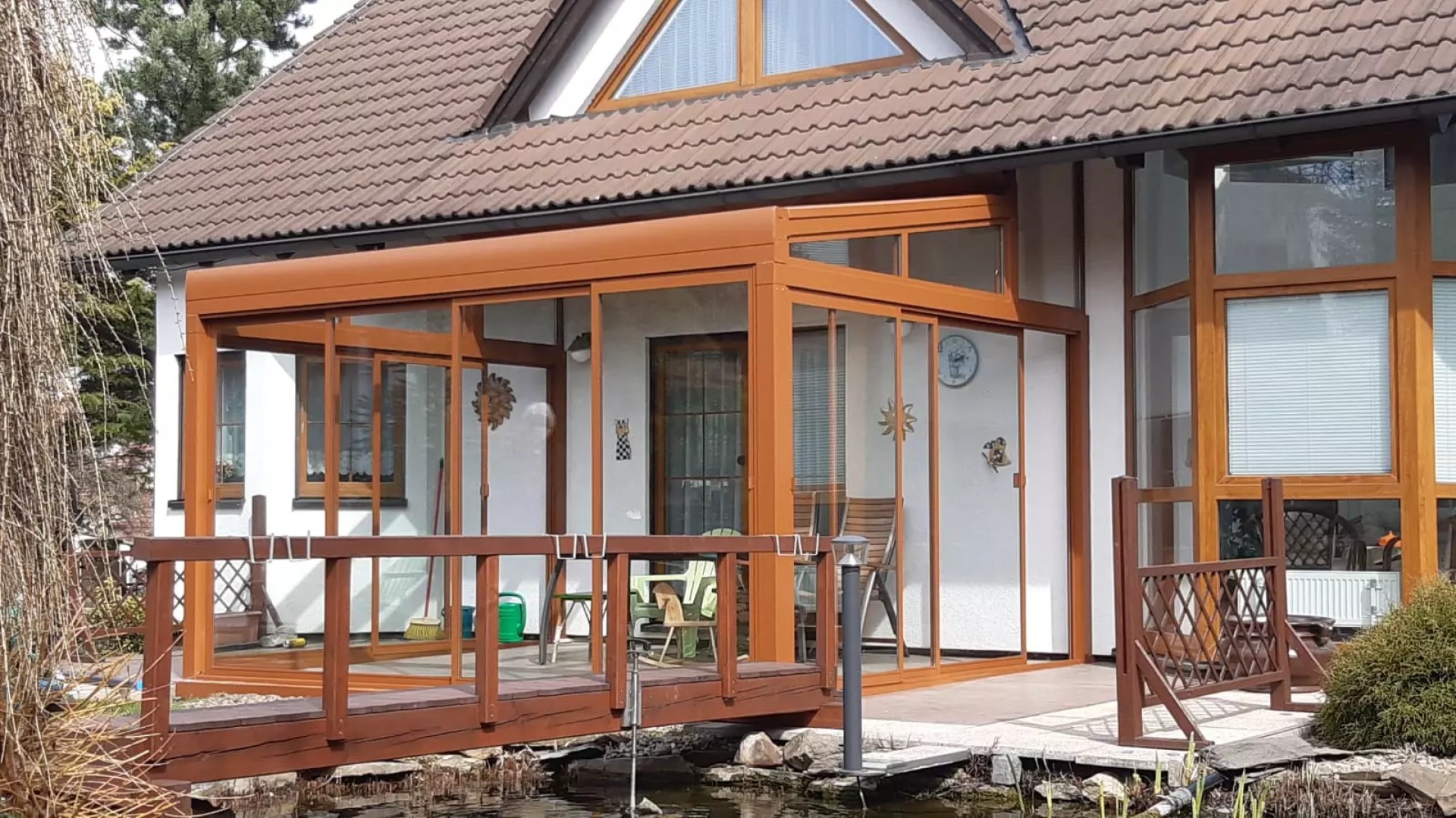 A brown ROBUST glass-enclosed aluminum pergola next to a single-family home with a wooden bridge