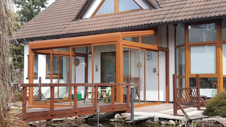 A brown ROBUST glass-enclosed aluminum pergola next to a single-family home with a wooden bridge
