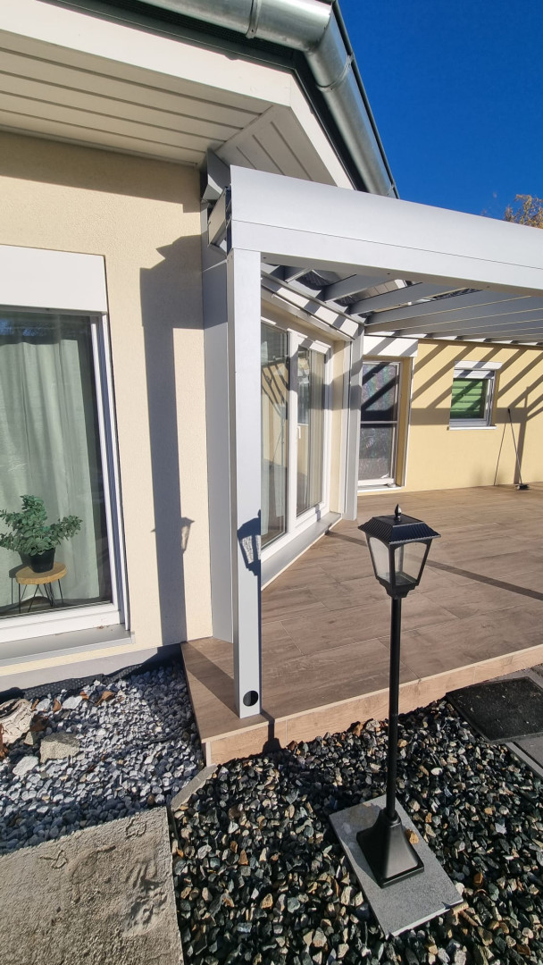 A close-up of how the white aluminum pergola is attached to the house's facade on a terrace with wooden trim.