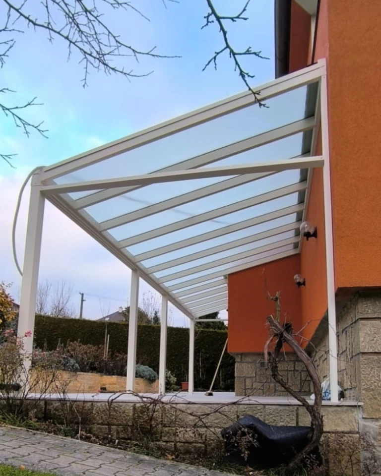 A modern white aluminum pergola with a glass roof attached to an orange single-family home
