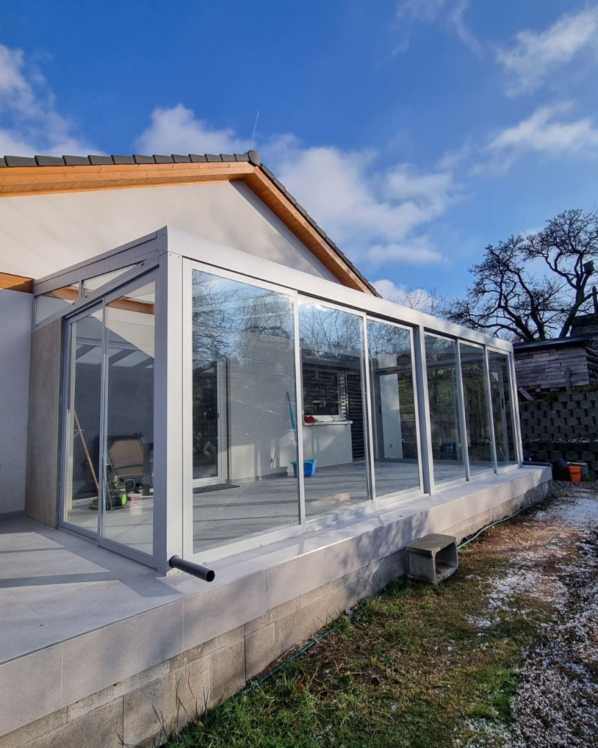 A modern, glass-enclosed sunroom with an aluminum frame attached to a single-family home