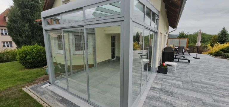 A modern, glass-enclosed sunroom with an aluminum frame, built on the spacious terrace of a single-family home.