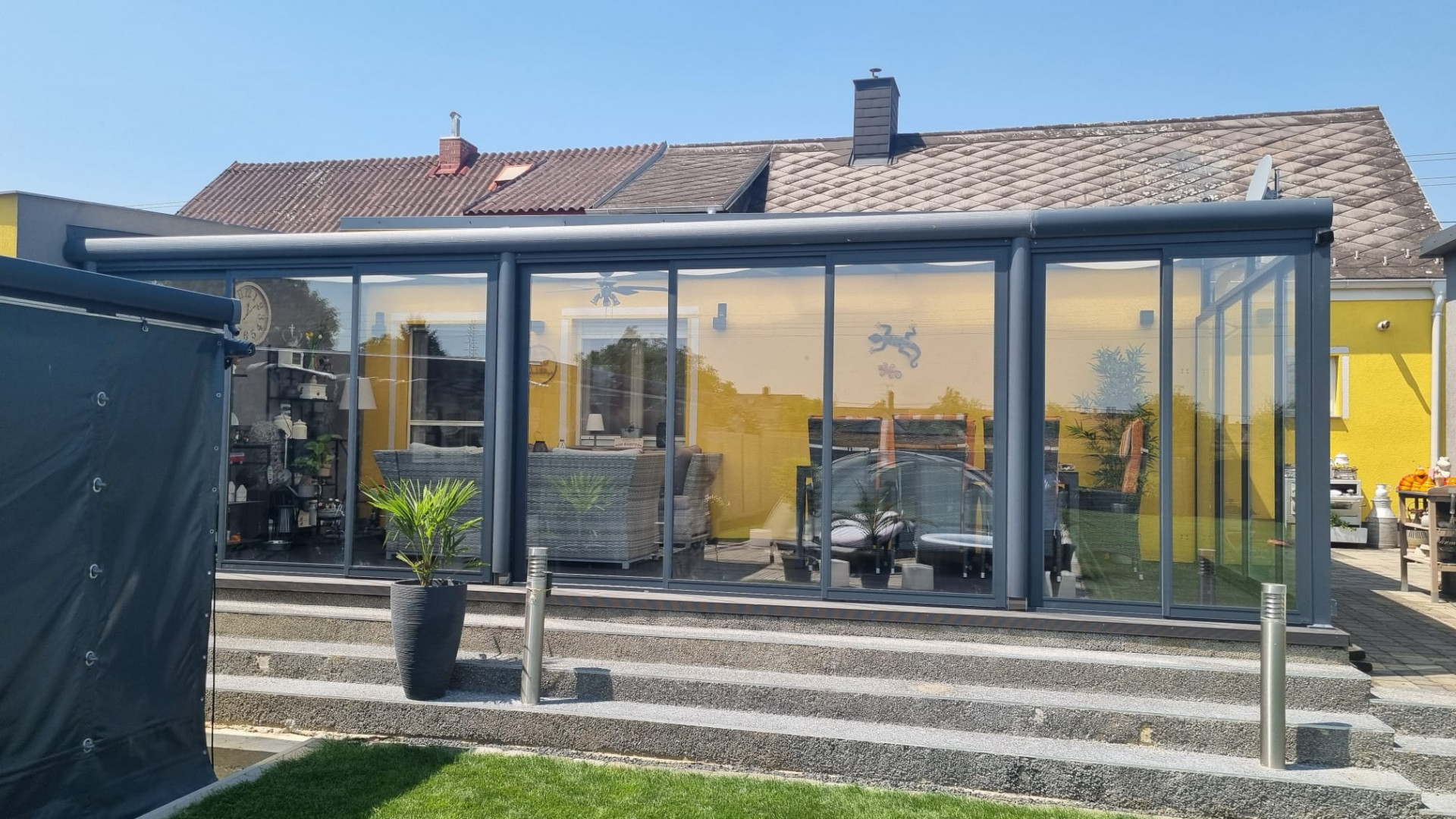 A modern, glass-enclosed aluminum sunroom in anthracite, attached to a yellow single-family home with a staircase.